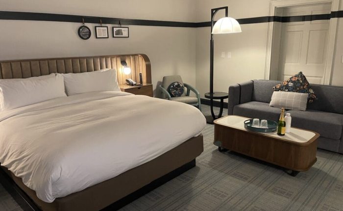 A cozy hotel room in one of the top hotels in Frederick MD, featuring a large bed with a gray headboard, white bedding, a gray armchair, sofa with patterned pillows, wooden coffee table with drinks, wall art above the bed, and a floor lamp.