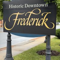 Outdoor sign reading 'Historic Downtown Frederick' in gold cursive on a dark panel, with greenery and street scene in background.