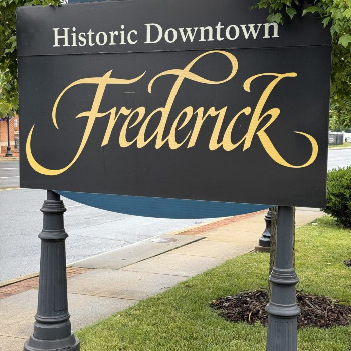 Outdoor sign reading 'Historic Downtown Frederick' in gold cursive on a dark panel, with greenery and street scene in background.