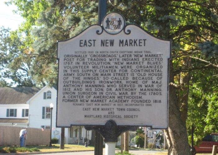 A historical marker for East New Market, MD, stands by a street. The sign details the town’s origins, role in the American Revolution and War of 1812, and mentions Dr. Anthony Manning. Trees and a house are visible in the background on a sunny day.