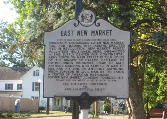 A historical marker for East New Market, MD, stands by a street. The sign details the town’s origins, role in the American Revolution and War of 1812, and mentions Dr. Anthony Manning. Trees and a house are visible in the background on a sunny day.
