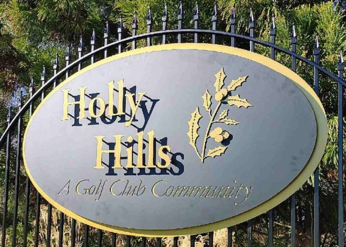 Oval sign reading Holly Hills: A Golf Club Community, with a holly branch graphic, mounted on a black wrought-iron fence