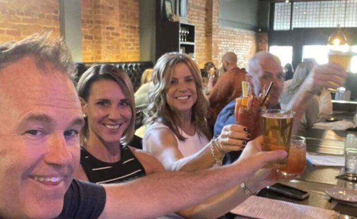 Four smiling adults sit at a restaurant table with exposed brick walls, raising drinks in a toast. Menus and a phone with a red case rest on the table, surrounded by the warm lighting and lively atmosphere of one of the top restaurants in Frederick MD.