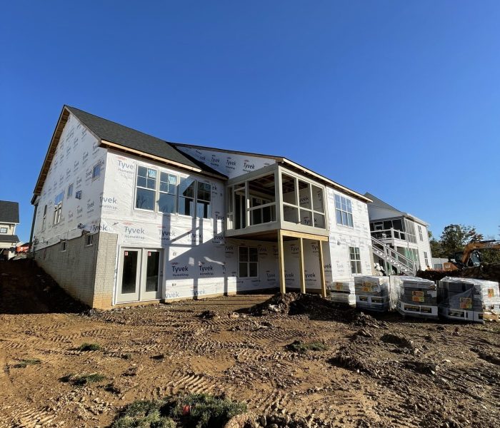 Two‑story house under construction with Tyvek wrap, exposed framing, and a dirt lot in the foreground while the sky is clear blue.