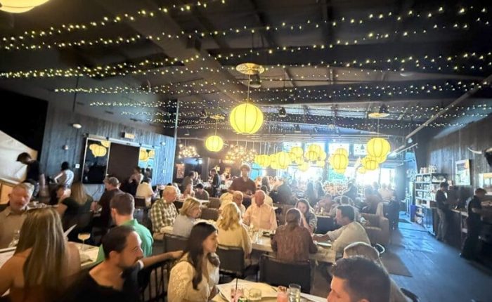 A lively restaurant scene with many people dining at tables under warm hanging lights and string lights on the ceiling. The room is bustling with conversation, and menus and drinks are visible on the tables. The atmosphere appears cozy and inviting.