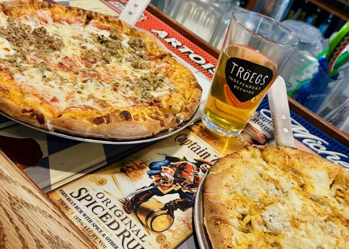 Two pizzas sit on metal trays atop a bar counter in pizza in Fredrick MD. One features sausage and cheese, the other is a garlicky white pizza. A pint of Tr&ouml;egs beer sits between them, with bar mats and drink bottles visible in the background.