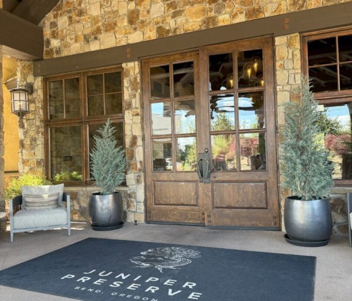 A stone building entrance with double wooden doors, flanked by large potted evergreens, welcomes visitors to Pronghorn Golf Course. A black mat in front reads JUNIPER PRESERVE, BEND, OREGON. Nearby sits a grey cushioned chair and lantern-style wall light.