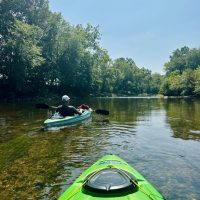 Two people kayak down a calm, shallow river near Mt Airy MD, surrounded by lush green trees under a clear blue sky. The photo is taken from one kayak looking ahead at the other, with sunlight reflecting off the water’s surface.