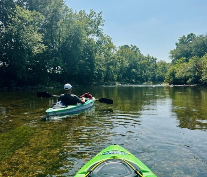 Two people kayak down a calm, shallow river near Mt Airy MD, surrounded by lush green trees under a clear blue sky. The photo is taken from one kayak looking ahead at the other, with sunlight reflecting off the water’s surface.