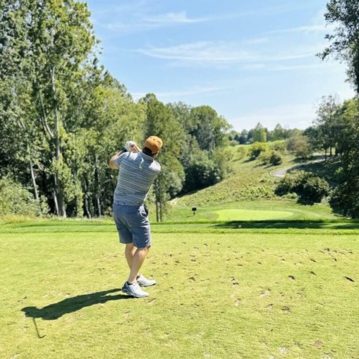 A man in a striped shirt, shorts, sneakers, and an orange cap swings a golf club on a green course surrounded by tall trees and rolling hills. Enjoy scenes like this at golf courses near Frederick, MD under the clear blue sky.