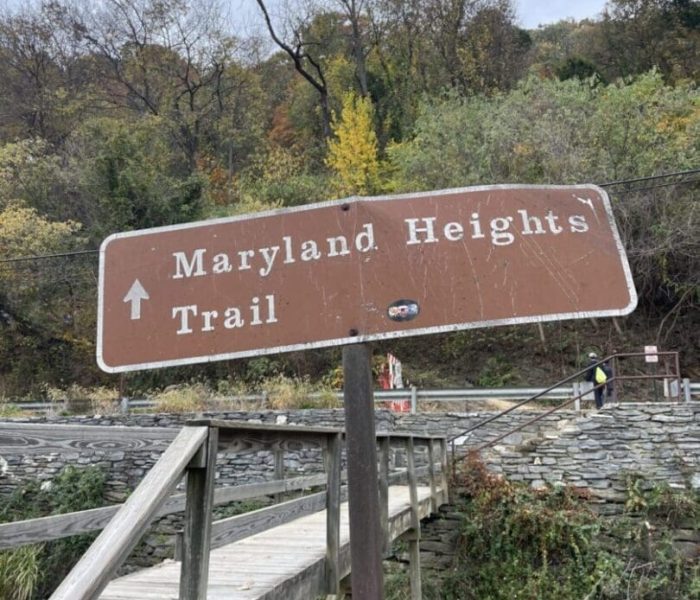 A brown wooden sign reads “Maryland Heights Trail” with a white arrow pointing up. The sign stands beside a wooden footbridge—one of the best hikes around Frederick MD—framed by stone walls and autumn trees under a cloudy sky.