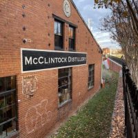 A red brick building in Frederick, MD with a black-and-white &ldquo;McClintock Distillery&rdquo; sign. Barred windows, a rooftop clock, graffiti, and autumn leaves by a black metal fence make it one of the unique things to do in Frederick. The sky is partly cloudy.