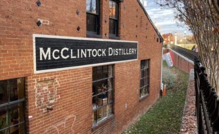 A red brick building in Frederick, MD with a black-and-white “McClintock Distillery” sign. Barred windows, a rooftop clock, graffiti, and autumn leaves by a black metal fence make it one of the unique things to do in Frederick. The sky is partly cloudy.