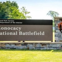 Monocacy-National-Battlefield-Frederick A large stone and metal sign on grass reads Monocacy National Battlefield, a top highlight among things to do in Frederick, MD. The National Park Service emblem is featured, with trees and a white building with a green roof in the background.