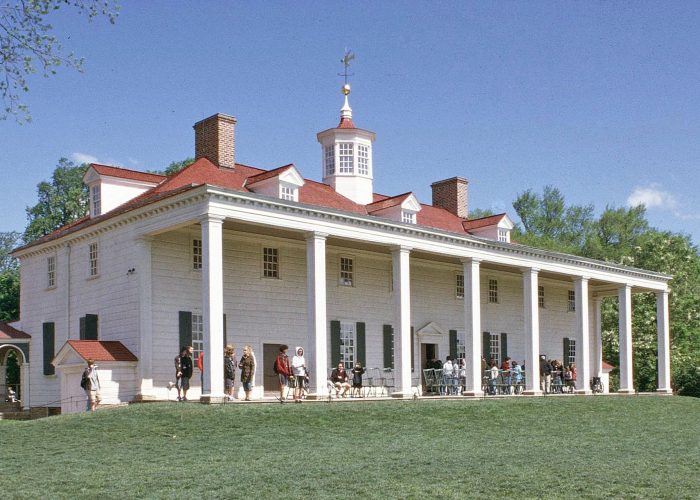 White two-story colonial house with a red roof and tall columns; people gathered on the veranda and lawn on a sunny day.