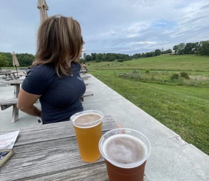 Nicole-at-Freys-Brewery-Mount-Airy A woman with shoulder-length hair sits at a wooden picnic table outdoors in New Market, MD, gazing toward a green field under a cloudy sky. Two plastic cups of beer rest on the table, with umbrellas and more tables visible in the background.