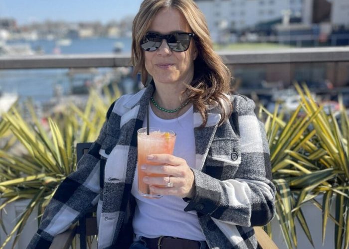 A woman wearing sunglasses and a black-and-white plaid jacket sits outdoors at a table in Annapolis, holding a pink drink with ice. She smiles slightly, with a sunny marina and buildings in the background—one of many relaxing things to do in Maryland.