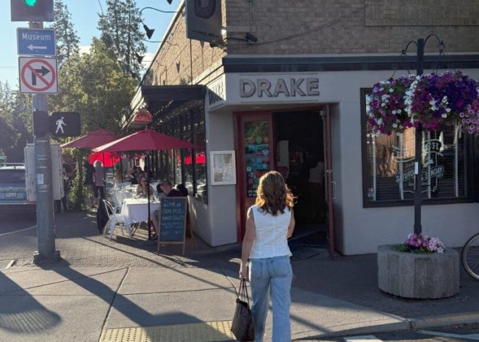 A woman in a white top and light blue jeans walks toward the entrance of Drake restaurant in downtown Bend, Oregon on a sunny day. Outdoor diners sit under red umbrellas as she explores Airbnb Bend Oregon nearby. Hanging flower baskets and a green traffic light are visible.