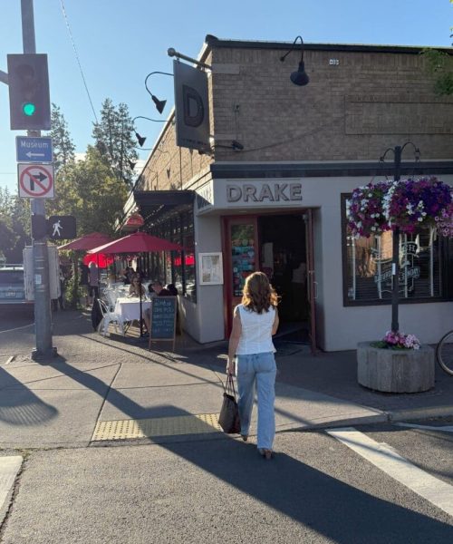A woman in a white top and light blue jeans walks toward the entrance of Drake restaurant in downtown Bend, Oregon on a sunny day. Outdoor diners sit under red umbrellas as she explores Airbnb Bend Oregon nearby. Hanging flower baskets and a green traffic light are visible.