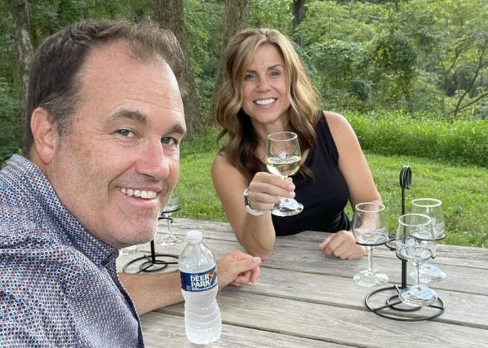 A man and woman sit at a wooden picnic table outdoors at Market Table Bistro, smiling at the camera. The woman holds a wine glass, with more empty glasses on the table. The man holds a water bottle amid lush green trees and grass on a sunny day.
