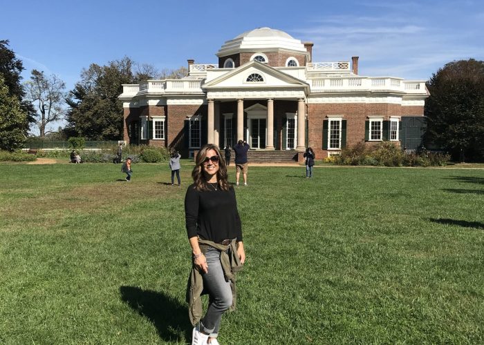 Woman in black long-sleeve top and jeans posing on a grassy lawn in front of a large brick neoclassical building with columns and a dome, under a clear blue sky.