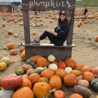 Nicole-pumpkin-patch-gaver-farm A woman with wavy brown hair, wearing sunglasses, a black outfit, and purple shoes, sits smiling in a wooden frame labeled pumpkins at Gaver Farm. She is surrounded by orange and multicolored pumpkins at a lively pumpkin patch.