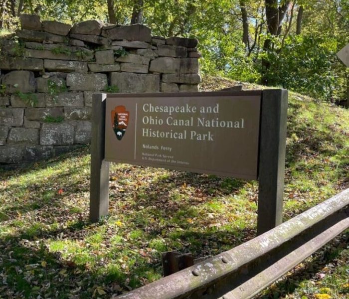A brown sign for Chesapeake and Ohio Canal National Historical Park stands amid grass and fallen leaves, near a wooden guardrail and stacked stone ruins, with tall, leafy trees casting dappled sunlight—perfect for exploring bike trails near Frederick MD.