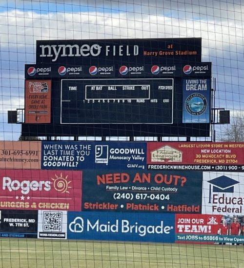 A baseball stadium scoreboard at Nymeo Field displays Pepsi logos and is blank, showing no scores or stats. Colorful advertisements for local businesses and the Frederick Keys schedule cover the lower part of the scoreboard, with netting in front.