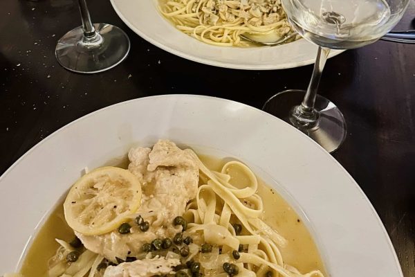 Two plates of pasta on a dark table at Cucina Massi Frederick MD: front dish features linguine with lemon, chicken, capers, and sauce; back dish has linguine with clams. Two glasses of white wine and a glass of water are also visible.