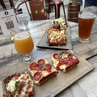 Two wooden boards hold Detroit-style pizza slices with toppings like pepperoni, sausage, jalape&ntilde;o, and vegetables. Nearby, two beers and a QR code menu add to the vibe of enjoying pizza in Fredrick MD at this inviting outdoor dining spot.