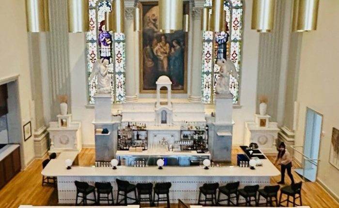 A stylish bar set inside a former church, this unique gem among restaurants in Frederick MD features high stained-glass windows, neoclassical columns, gold pendant lights, a marble bar lined with bottles, black barstools, and round tables on a wooden floor.