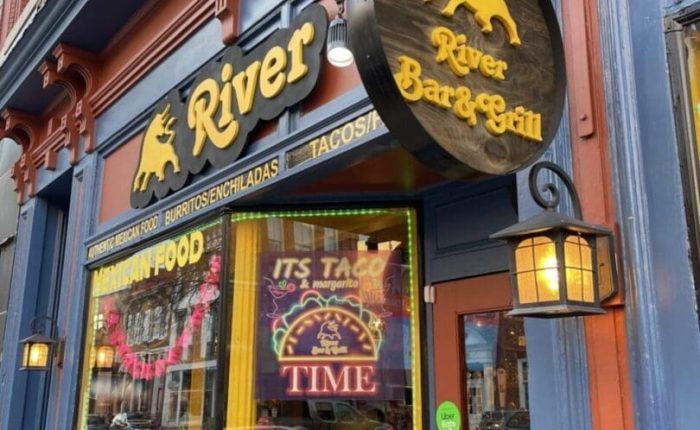 The entrance of River Bar & Grill, one of the best bars in Frederick, features a round wooden sign with a golden bull and yellow lettering. The blue and orange exterior boasts MEXICAN FOOD and ITS TACO TIME signs, decorative lanterns, and brick details.