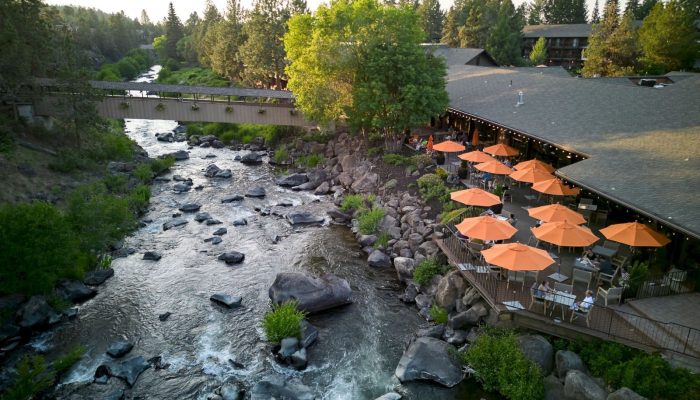 A riverside restaurant patio with many orange umbrellas, built on a wooden deck beside a rocky, flowing river and a pedestrian bridge nearby.