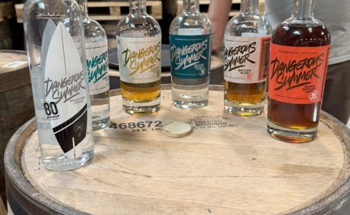 Six bottles of Dangerous Summer spirits, featured at some of the best bars in Frederick, sit on a wooden barrel with vibrant labels. Barrels fill the background while a person in a gray shirt stands behind them, arms partially visible.