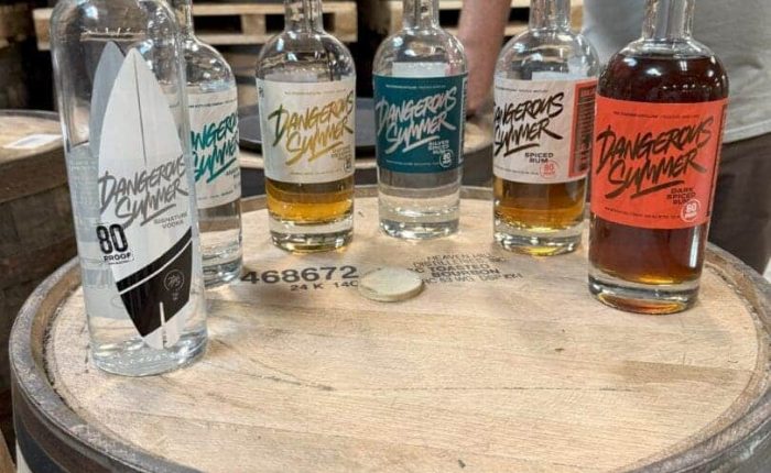 Six bottles of Dangerous Summer spirits, featured at some of the best bars in Frederick, sit on a wooden barrel with vibrant labels. Barrels fill the background while a person in a gray shirt stands behind them, arms partially visible.