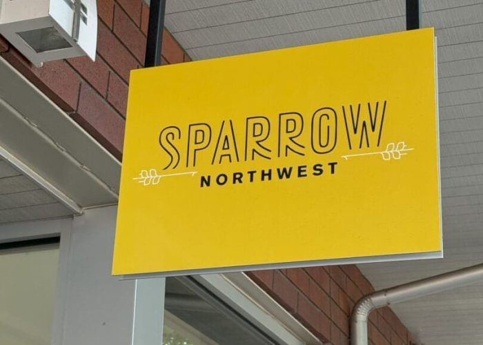 A yellow hanging sign with “Sparrow Northwest” in bold black letters and simple decorative lines, mounted under a covered walkway with a red brick wall and windows in the background.