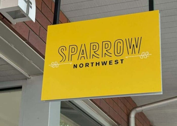 A yellow hanging sign with “Sparrow Northwest” in bold black letters and simple decorative lines, mounted under a covered walkway with a red brick wall and windows in the background.