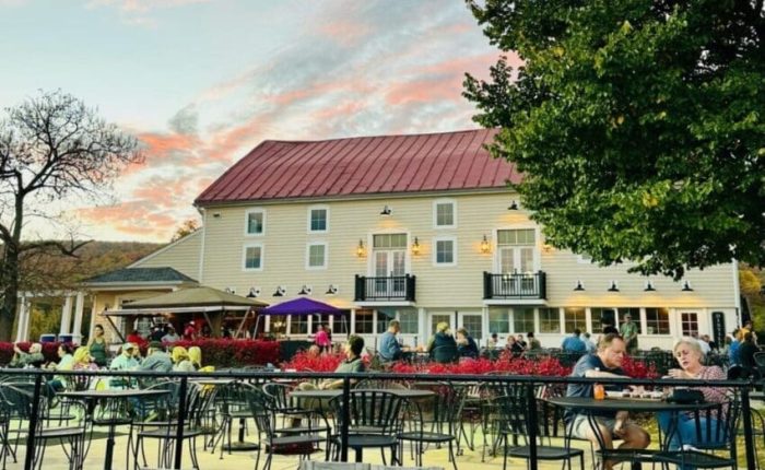 An outdoor dining area with wooden and metal tables and chairs is filled with people socializing. Behind them stands a large beige building, similar to some hotels in Frederick MD, under a colorful sunset sky framed by green trees.