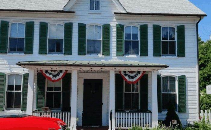 A white two-story house with green shutters, a metal porch roof, and patriotic red, white, and blue bunting above the entrance evokes classic charm in New Market, MD. A red car is partially visible by shrubs and a white picket fence lining the porch.