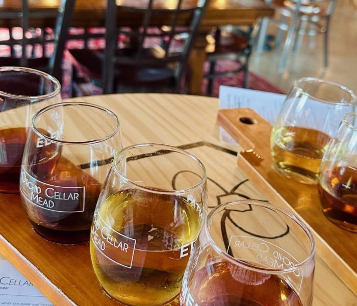 A wooden table at Orchid Cellar Middletown MD holds a flight of four glasses filled with different colored meads, ranging from amber to golden. The glasses sit on a labeled tray, with empty chairs and a brick wall in the cozy tasting room.