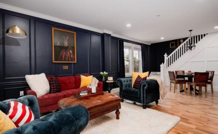 A stylish living room inspired by boutique hotels in downtown Frederick MD, with dark navy paneled walls, a red sofa, green velvet chairs, and a whimsical rabbit portrait above. Hardwood floors lead to a dining area and white staircase in the background.