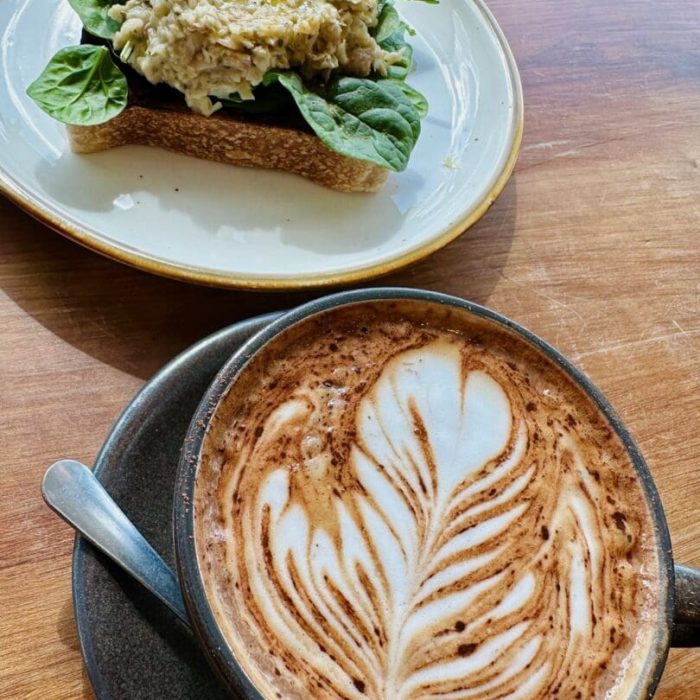 A cup of cappuccino with leaf-shaped latte art sits beside an open-faced sandwich topped with spinach and a creamy spread, all on a wooden table at a cozy coffee shop in Frederick MD.