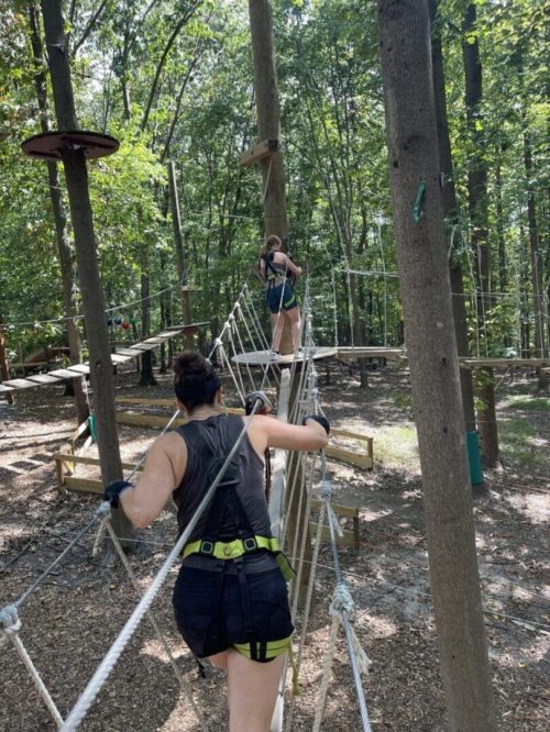 Two people wearing harnesses and gloves cross a rope bridge in a forested adventure park near New Market MD. Sunlight filters through the trees onto the woodchip-covered ground as one person grips the ropes, while the other moves confidently ahead.