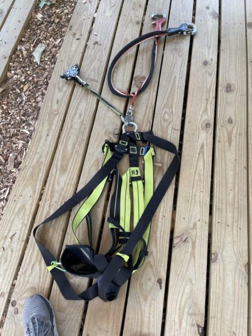 Tree Trekkers Harness A yellow and black safety harness with attached lanyard and metal clips is laid out on a wooden deck in New Market, MD. The harness is loosened and unbuckled, spread out flat. A person’s gray shoe is visible at the lower left corner of the image.