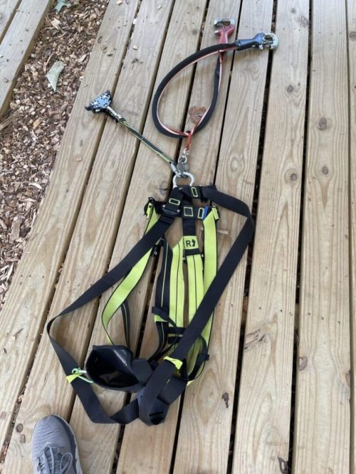 A yellow and black safety harness with attached lanyard and metal clips is laid out on a wooden deck in New Market, MD. The harness is loosened and unbuckled, spread out flat. A person’s gray shoe is visible at the lower left corner of the image.