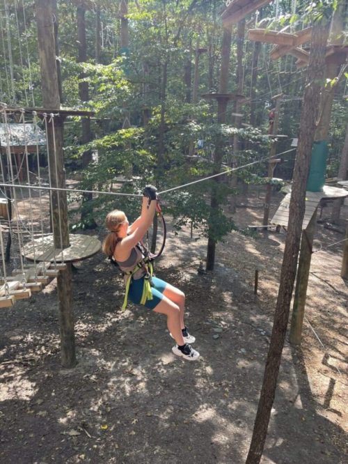 Tree Trekkers Zipline A person with long hair in a ponytail, wearing a harness, helmet, blue shorts, and sneakers, is ziplining through a forest adventure park in New Market MD, with tall trees and sunlight filtering through leaves onto ropes course structures.