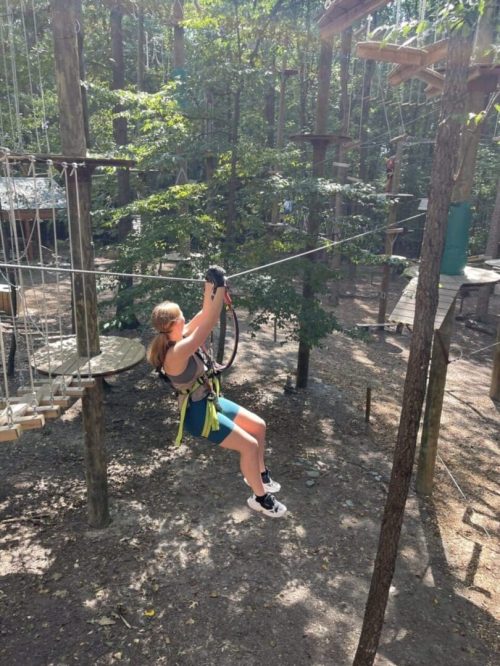 A person with long hair in a ponytail, wearing a harness, helmet, blue shorts, and sneakers, is ziplining through a forest adventure park in New Market MD, with tall trees and sunlight filtering through leaves onto ropes course structures.