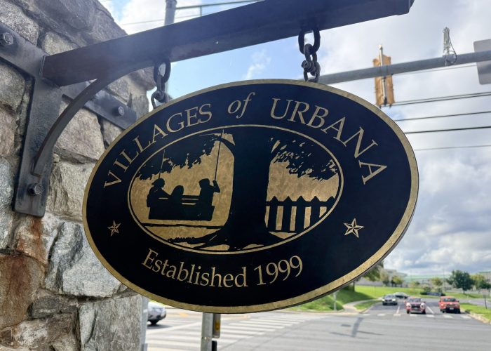 Round black circular sign with a gold border hanging from a stone wall; features a world map and the text 'Established 1999'.