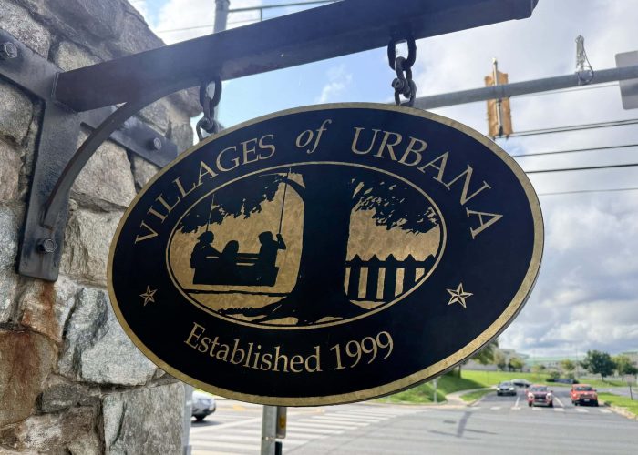 Round black circular sign with a gold border hanging from a stone wall; features a world map and the text 'Established 1999'.