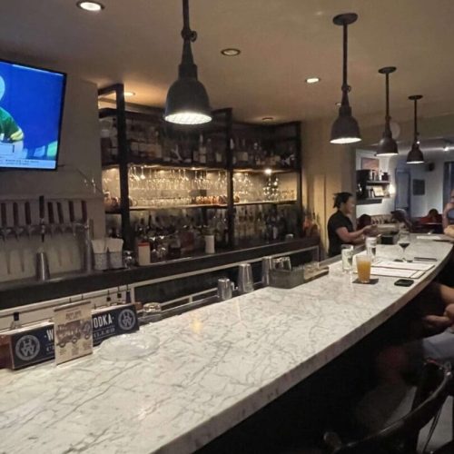 A marble-topped bar with hanging pendant lights, shelves stocked with liquor bottles and glassware behind it in New Market MD. A TV shows a sports event as several people sit at the bar, one with a drink, in a dimly lit, cozy atmosphere.